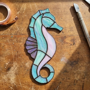 Puede incluir: Un caballito de mar de vidriera en tonos azules, turquesas y lavanda. El caballito de mar está delineado en negro y se encuentra sobre una superficie de madera. Es un objeto decorativo.