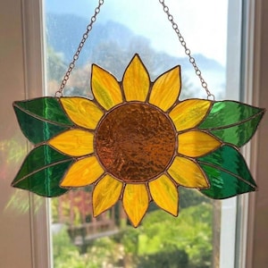 May include: A stained glass sunflower suncatcher with yellow petals, a brown centre, and green leaves. The decorative piece is suspended by a chain and hangs in front of a window, showcasing the vibrant colours.