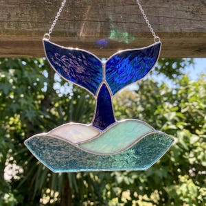 May include: Stained glass whale tail suncatcher with shades of blue and green. The whale tail is a deep blue with lighter blue accents. The waves below are shades of green and white. A silver chain is attached for hanging.