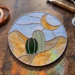 Op de afbeelding: Rond gebrandschilderd glaskunstwerk met een woestijnlandschap met een groene cactus, een gele halve maan en een blauwe lucht. Het glas is omlijst met zilver soldeer en heeft een diameter van ongeveer 20 cm.