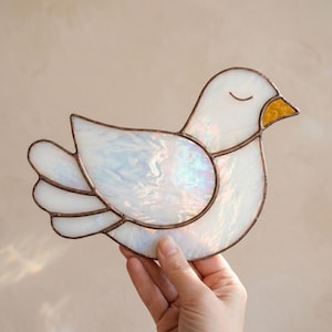 May include: A stained glass dove with a white body and iridescent wings. The bird has a yellow beak and closed eyes, outlined with a copper-colored metal. The dove is a decorative item.