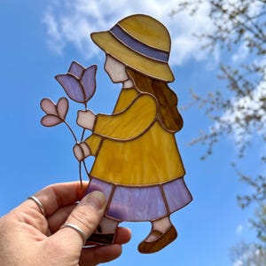 Op de afbeelding: Glazen figuur van een meisje in een gele jurk en hoed, met paarse en roze bloemen. De figuur heeft bruine schoenen en haar, met een blauwe lucht op de achtergrond. Het glas is omlijnd in donkerbruin.