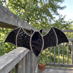 Puede incluir: Un atrapasoles de vidrieras con forma de murciélago, compuesto por paneles de vidrio negro texturizado unidos por líneas de soldadura de metal plateado. El murciélago tiene orejas puntiagudas y alas extendidas, suspendido de una cadena metálica.
