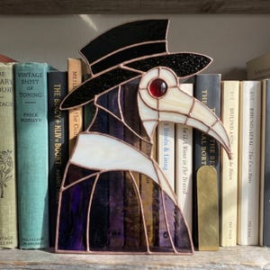 May include: A stained glass plague doctor figure featuring a black top hat, a cream-coloured beak mask with a single red circular eye, and a purple robe. The piece is constructed with dark metal solder lines and stands upright as a decorative object.