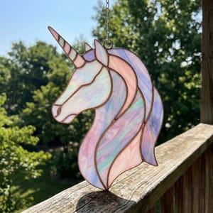 May include: A stained glass unicorn head with a pink, purple, and white color scheme. The unicorn has a long, spiraled horn and is hanging from a chain. The glass is iridescent, reflecting light. The piece is set against a backdrop of green trees.