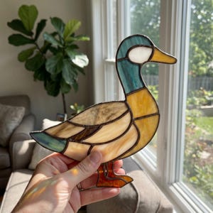 Puede incluir: Figura de pato de vidriera con cabeza turquesa, cuerpo amarillo y alas marrones. El pato tiene pico y patas naranjas. El vidrio está segmentado con líneas oscuras, creando un efecto de mosaico.