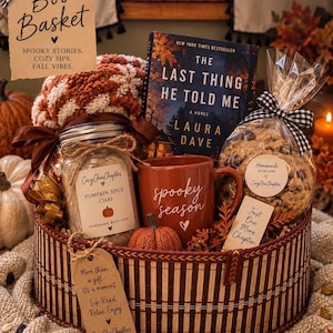 May include: A round woven basket containing a book titled The Last Thing He Told Me, a red mug with the text spooky season, a jar of pumpkin spice chai, a bag of cookies with a black and white gingham bow, and a tag reading Boo Basket.
