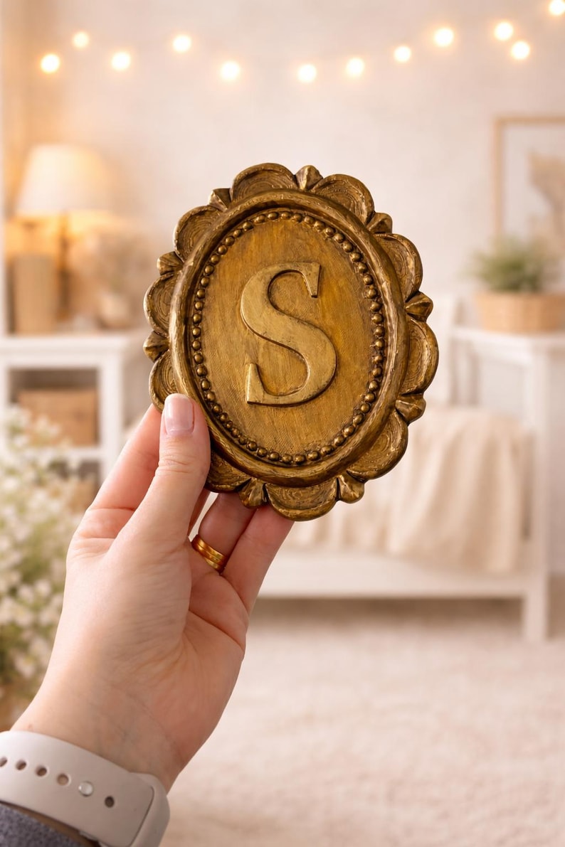 May include: An oval-shaped gold-colored plaque featuring a raised letter S in the center. The plaque has a decorative scalloped edge with a beaded border detail surrounding the central letter. The finish appears textured with a metallic sheen.
