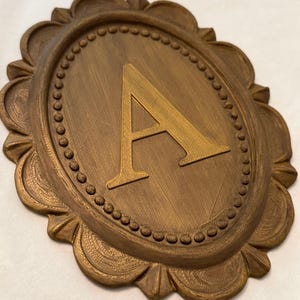May include: An oval-shaped wall plaque featuring a raised letter A in the center. The plaque has a bronze-colored finish with a textured surface. The border consists of a beaded inner ring surrounded by a scalloped edge with fine line details.