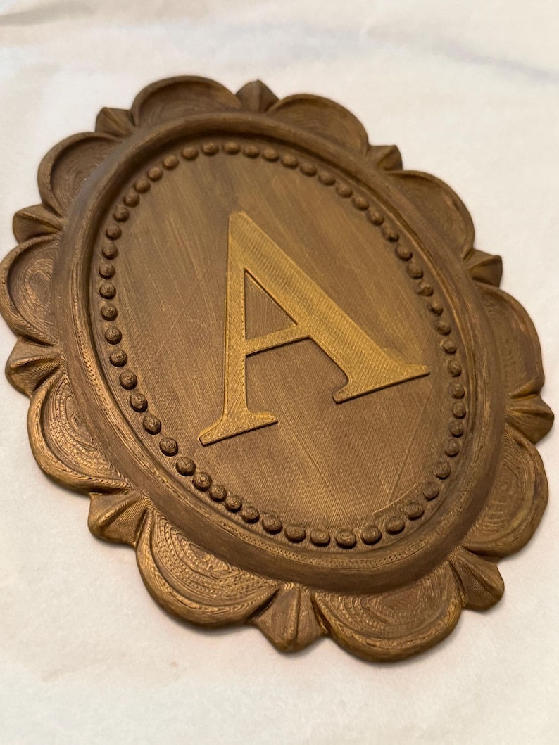 May include: An oval-shaped wall plaque featuring a raised letter A in the center. The plaque has a bronze-colored finish with a textured surface. The border consists of a beaded inner ring surrounded by a scalloped edge with fine line details.