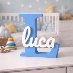 Puede incluir: Una letra "L" azul de madera con el nombre "Luca" en escritura blanca. La decoración de la guardería incluye una cuna, una pila de anillos de colores y una pared azul claro con decoraciones de estrellas.