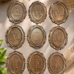 May include: Nine oval-shaped plaques arranged in a grid, each featuring a raised capital letter in the center. The letters displayed are B, K, M, M, O, M, T, E, and L. Each plaque has a dark bronze finish with a beaded border and scalloped edges.