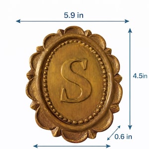 May include: An oval gold-colored plaque featuring a raised letter S in the center, surrounded by a beaded border and a scalloped edge. The plaque has approximate measurements of 5.9 inches in width, 4.5 inches in height, and 0.6 inches in depth.
