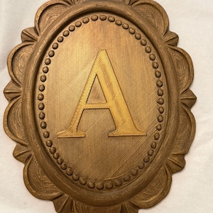 May include: An oval-shaped wall plaque in a metallic bronze finish featuring a raised capital letter A in the center. The plaque is surrounded by a decorative border with a beaded inner ring and a scalloped outer edge with textured details.