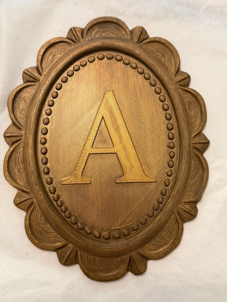 May include: An oval-shaped wall plaque in a metallic bronze finish featuring a raised capital letter A in the center. The plaque is surrounded by a decorative border with a beaded inner ring and a scalloped outer edge with textured details.