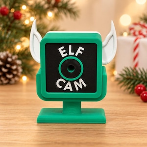 May include: A green square-shaped camera prop with white pointed elf ears on the sides. The front face is black with a central circular lens opening and the white text ELF CAM printed above and below the lens. It stands on a flat green base.