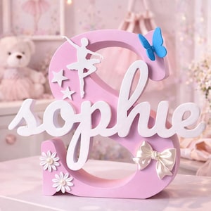 Può includere: Una lettera "S" rosa in legno con il nome "Sophie" in corsivo bianco. Presenta una ballerina, stelle, una farfalla blu, un fiocco e decorazioni floreali. Un oggetto decorativo per la cameretta di un bambino.