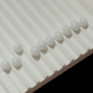 Puede incluir: Una colección de pequeñas cuentas esféricas blancas, opacas y lisas, dispuestas sobre una superficie blanca que presenta una serie de crestas paralelas en relieve. Las cuentas están dispersas por la superficie texturizada, creando un contraste entre las formas redondas y el patrón lineal de la base.