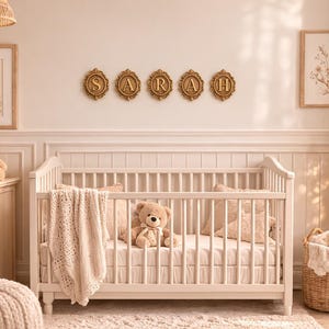 May include: A white wooden crib with a cream-colored knit blanket and a plush teddy bear inside. Above the crib, five gold-toned oval frames display the letters S, A, R, A, and H. The room features neutral tones, teddy bear decor, and floral art.