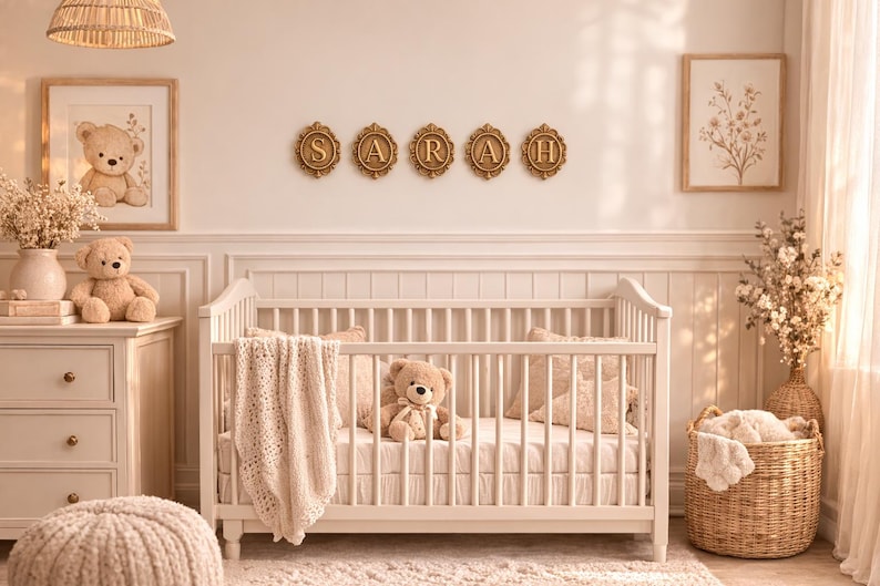 May include: A white wooden crib with a cream-colored knit blanket and a plush teddy bear inside. Above the crib, five gold-toned oval frames display the letters S, A, R, A, and H. The room features neutral tones, teddy bear decor, and floral art.