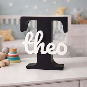 Pode incluir: Uma letra "T" de madeira preta com o nome "theo" em branco. A peça decorativa é exibida em uma cômoda branca em um ambiente de berçário. O fundo inclui um berço e iluminação suave.