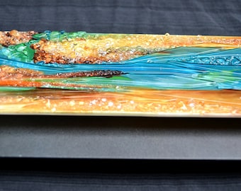May include: A rectangular resin art piece depicting a landscape with a river. The artwork features layers of orange, yellow, green, and blue resin, creating a textured, three-dimensional effect. The river is a vibrant blue.