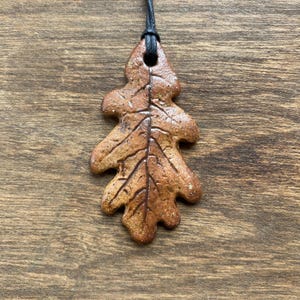 May include: A brown ceramic pendant shaped like an oak leaf with a textured, speckled surface and dark brown vein details. The pendant features a hole at the top for a black cord, which is threaded through to form a necklace for wearing.