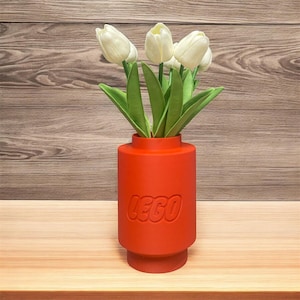 May include: A red vase in the style of a LEGO brick, embossed with the LEGO logo, containing white tulips and green leaves. The vase sits on a wooden surface against a wooden backdrop.