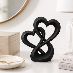 May include: A matte black sculpture featuring two intertwined heart shapes that form a continuous, fluid loop. The object has a textured surface and stands upright, showcasing a symmetrical design with open spaces between the curved heart forms.
