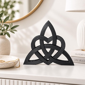 May include: A black wooden sculpture featuring an intricate triquetra knot design with three interconnected loops forming a central heart shape. The object has a smooth matte finish and stands upright on a flat surface, displaying a symmetrical pattern.