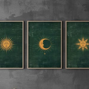 May include: A set of three framed wall art prints featuring gold celestial illustrations of a sun, a crescent moon, and a star on a dark forest green textured background. Each print is encased in a light wood frame with a clean, simple border.