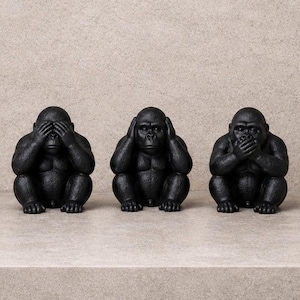 Puede incluir: Tres figuras de gorilas negros en la pose de "no ver el mal, no oír el mal, no hablar el mal". Las estatuas están sobre una estantería gris claro contra una pared beige claro texturizada. Los gorilas están sentados.