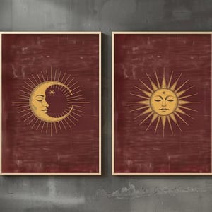 May include: Two framed rectangular wall art prints featuring a deep maroon background. The left print displays a golden crescent moon with a human face and radiating sunburst lines. The right print shows a golden sun with a human face and rays.