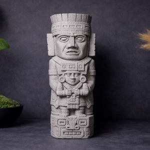 May include: A grey stone statue of an ancient figure with detailed carvings. The statue is flanked by a vase with green leaves, a black bowl of moss, and a vase with dried ornamental grass. The background is a dark grey.