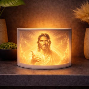 Può includere: Una luce notturna rettangolare bianca con un'immagine illuminata di Gesù Cristo con un'aureola radiosa. L'immagine è centrata nella luce, che è posta su una superficie scura. Vasi decorativi sono sullo sfondo.