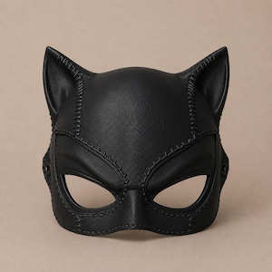 Op de afbeelding: Een zwart masker in de vorm van een kat met puntige oren en amandelvormige oogopeningen. Het masker is voorzien van zichtbare decoratieve stiksels langs de randen en over het voorhoofd, wat zorgt voor een gestructureerd uiterlijk met een gladde afwerking.