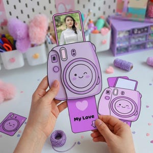 Puede incluir: Una manualidad de papel morado claro de una cámara con una foto de una persona. La cámara tiene una cara sonriente y las palabras "My Love" en la foto. Suministros de manualidades en el fondo.