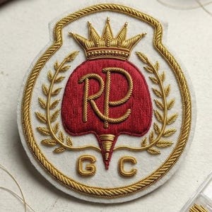 Könnte beinhalten: Ein kreisförmiger Stickaufnäher auf weißem Filz mit einer goldenen Krone über einem roten Schild mit den golden gestickten Buchstaben RP. Der Schild ist von goldenen Weizenähren umrahmt, mit den goldenen Buchstaben G und C darunter.