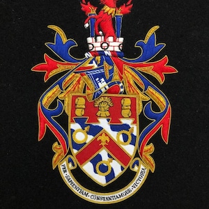 Puede incluir: Escudo bordado sobre tela negra. El escudo presenta un ciervo rojo sobre una torre blanca, con un escudo debajo. El escudo tiene un cheurón rojo sobre fondo blanco, con detalles azules y dorados. El texto "PER SAPIENTIAM CONSTANTIAMQUE VICTORIA" está arqueado debajo.