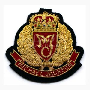 May include: Embroidered patch with a black background, gold laurel leaves, and a red shield bearing the initials "MJ". A gold crown sits atop the shield. The text "MICHAEL JACKSON" is displayed on a red banner.
