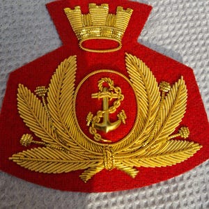 Peut inclure: Un écusson en feutre rouge avec une broderie métallique dorée. Le motif présente une ancre centrale entourée d'une corde, flanquée de deux grandes palmes et surmontée d'une couronne murale dorée. La broderie utilise du fil cannetille.