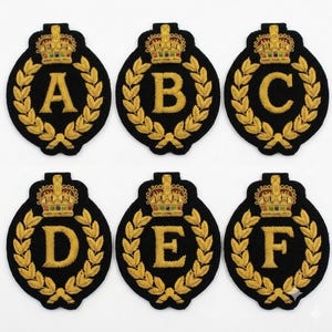 Könnte beinhalten: Sechs kreisförmige, schwarze Stickaufnäher mit einem goldenen Lorbeerkranzrand, einer goldenen Krone an der Oberseite und einem großen goldenen Buchstaben in der Mitte. Die Buchstaben A, B, C, D, E und F sind in zwei Reihen alphabetisch angeordnet.