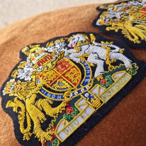 Può includere: Due toppe ricamate raffiguranti lo stemma reale del Regno Unito. Il design include un leone dorato e un unicorno argentato che fiancheggiano uno scudo centrale con dettagli rossi, oro e blu, il tutto cucito su una base di tessuto blu scuro.
