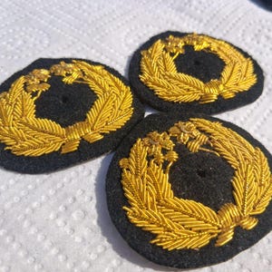 Op de afbeelding: Drie ronde zwarte stoffen patches met gedetailleerd borduurwerk van gouden metaaldraad. Elke patch toont een kransontwerp bestaande uit gestileerde bladeren en kleine bloemelementen, met een klein rond gaatje in het midden.