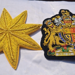 Op de afbeelding: Twee geborduurde patches. De ene is een gouden, achtpuntige ster gemaakt van metaaldraad. De andere is een zwarte, schildvormige patch met een gedetailleerd koninklijk wapenschild in goud-, zilver-, rood- en blauwdraad, inclusief de tekst DIEU ET MON DROIT.