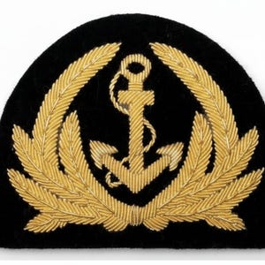 May include: A black, semi-circular patch featuring a gold anchor emblem surrounded by a gold wheat-like wreath. The anchor is centered, with the wreath's design mirroring on both sides. The patch has a velvet-like texture.