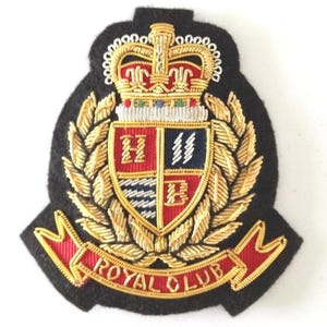 Könnte beinhalten: Gestickter Aufnäher mit heraldischem Design. Der Aufnäher zeigt eine Krone, einen in vier Abschnitte unterteilten Schild und ein Banner mit der Aufschrift "ROYAL CLUB". Der Aufnäher ist hauptsächlich schwarz, mit goldenen, roten, weißen und blauen Akzenten.