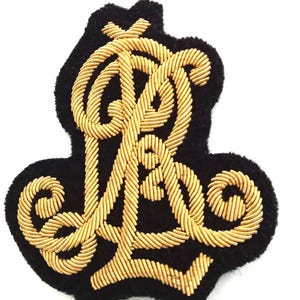 May include: A black embroidered patch with gold-colored thread. The patch features an ornate, intertwined design of the letters "R" and "L". The patch is likely for decorative purposes, such as on clothing or accessories.