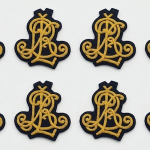 May include: Eight identical navy blue fabric patches arranged in two rows of four. Each patch features an intricate, gold-colored embroidered monogram consisting of the intertwined letters R and L, with decorative swirling flourishes at the base.
