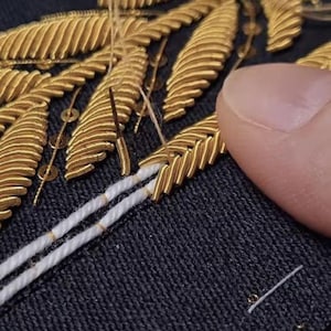 Puede incluir: Una superficie de tela oscura con un intrincado bordado metálico dorado en forma de hojas. El diseño incorpora pequeñas lentejuelas doradas y finas líneas de hilo blanco. Se ve una aguja cosiendo activamente los elementos metálicos a la tela.